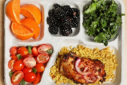 A lunch tray: orange slices, blackberries, kale salad, cherry tomato salad, and jollof rice and chicken.